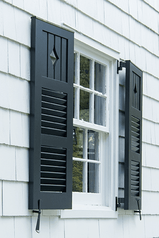 Pair of black louvered shutters held open on wrought iron dogs either side of a window in the white shingles of a Westfield, NJ exterior renovation