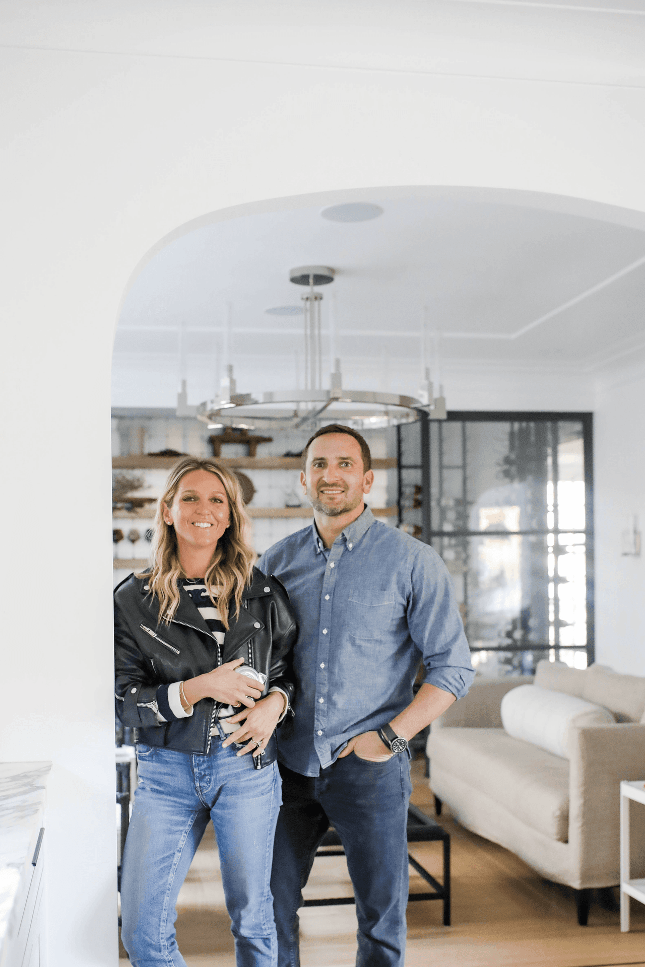 Mike and Ellie Mroz under an arched cased opening in a Westfield, NJ home, with a ring chandelier and steel-framed glass doors behind them