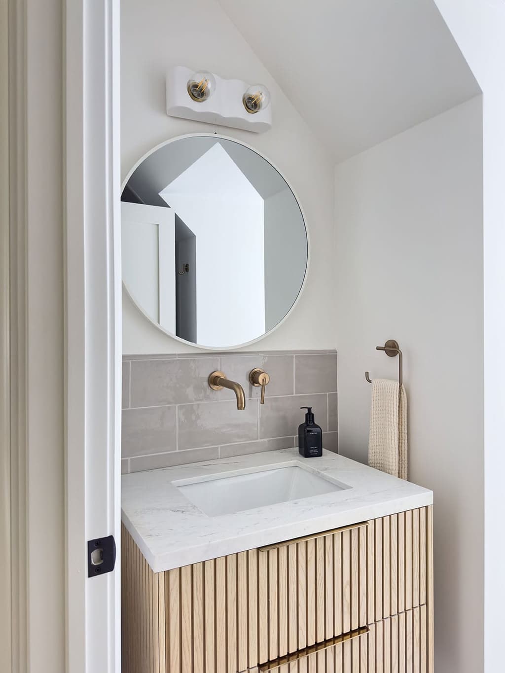 Powder room with a fluted oak vanity under a white stone counter, a brass wall-mounted faucet, a round mirror and a sconce above
