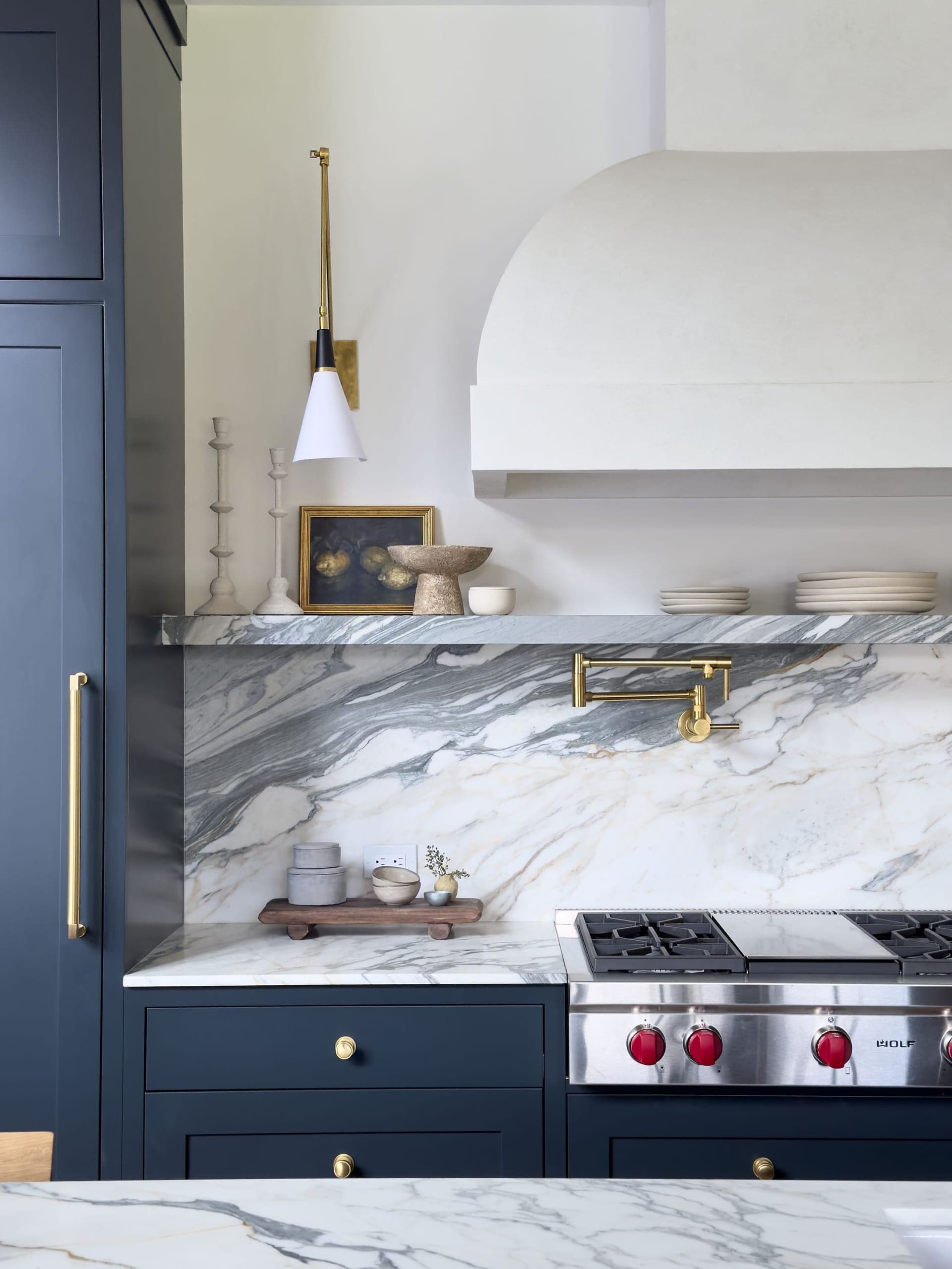 Kitchen range wall with a plaster hood, book-matched marble backsplash, navy cabinetry and a brass pot filler