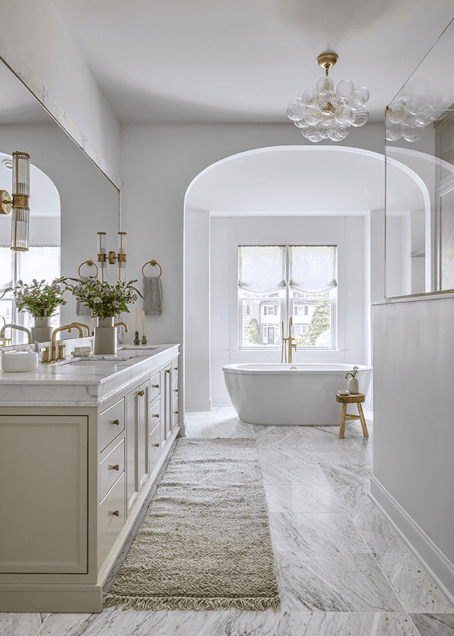 White bathroom in a Westfield, NJ new-construction home, with a long marble vanity, a crystal chandelier and a freestanding tub in an arched alcove at the end