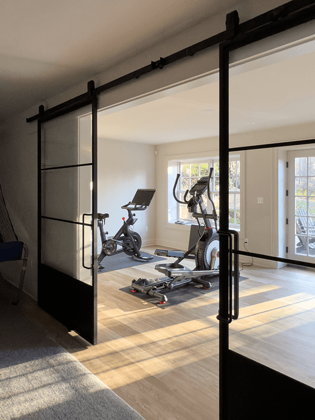 Black steel and glass sliding doors standing open on a home gym in a Summit, NJ renovation, with a bike and a cross-trainer by the windows