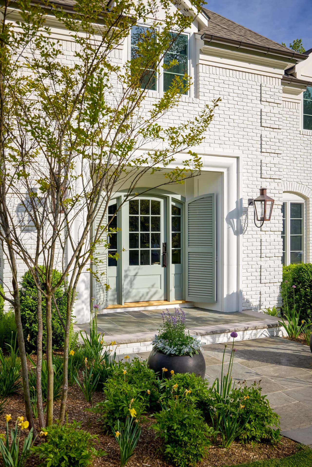 Arched entry of a Mountainside, NJ home renovation seen at an angle, with a blue-green door, a lantern on the painted brick and daffodils in the bed below
