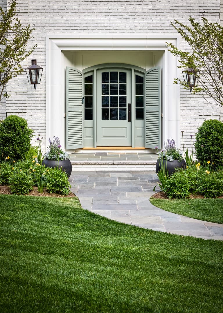 Sage-green arched front door with sidelights, louvered shutters and copper lanterns, set into painted white brick at a Mountainside, NJ exterior renovation by Michael Robert Construction