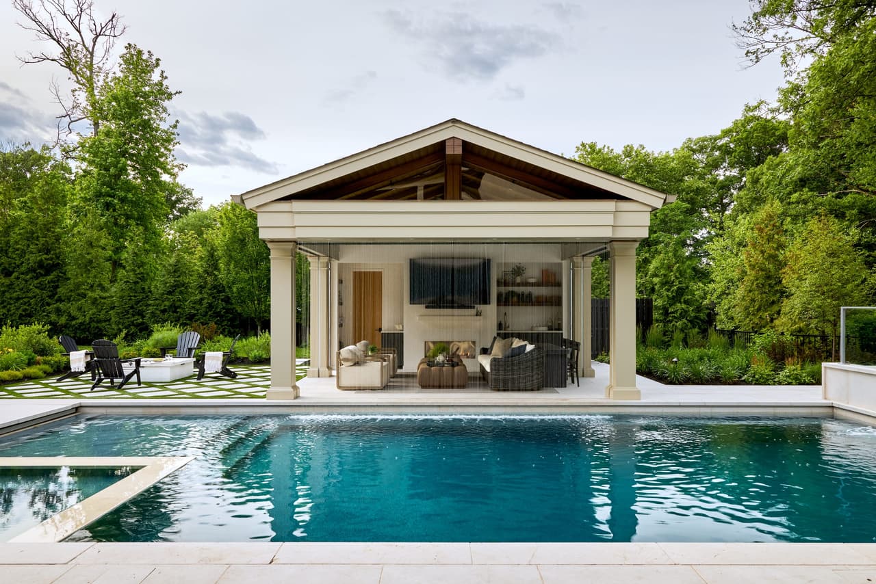 Gabled pool cabana seen across the pool at a Westfield, NJ home, with columned openings, an open-air lounge and a stone outdoor fireplace, built by Michael Robert Construction