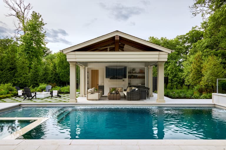 Gabled pool cabana seen across the pool at a Westfield, NJ home, with columned openings, an open-air lounge and a stone outdoor fireplace, built by Michael Robert Construction