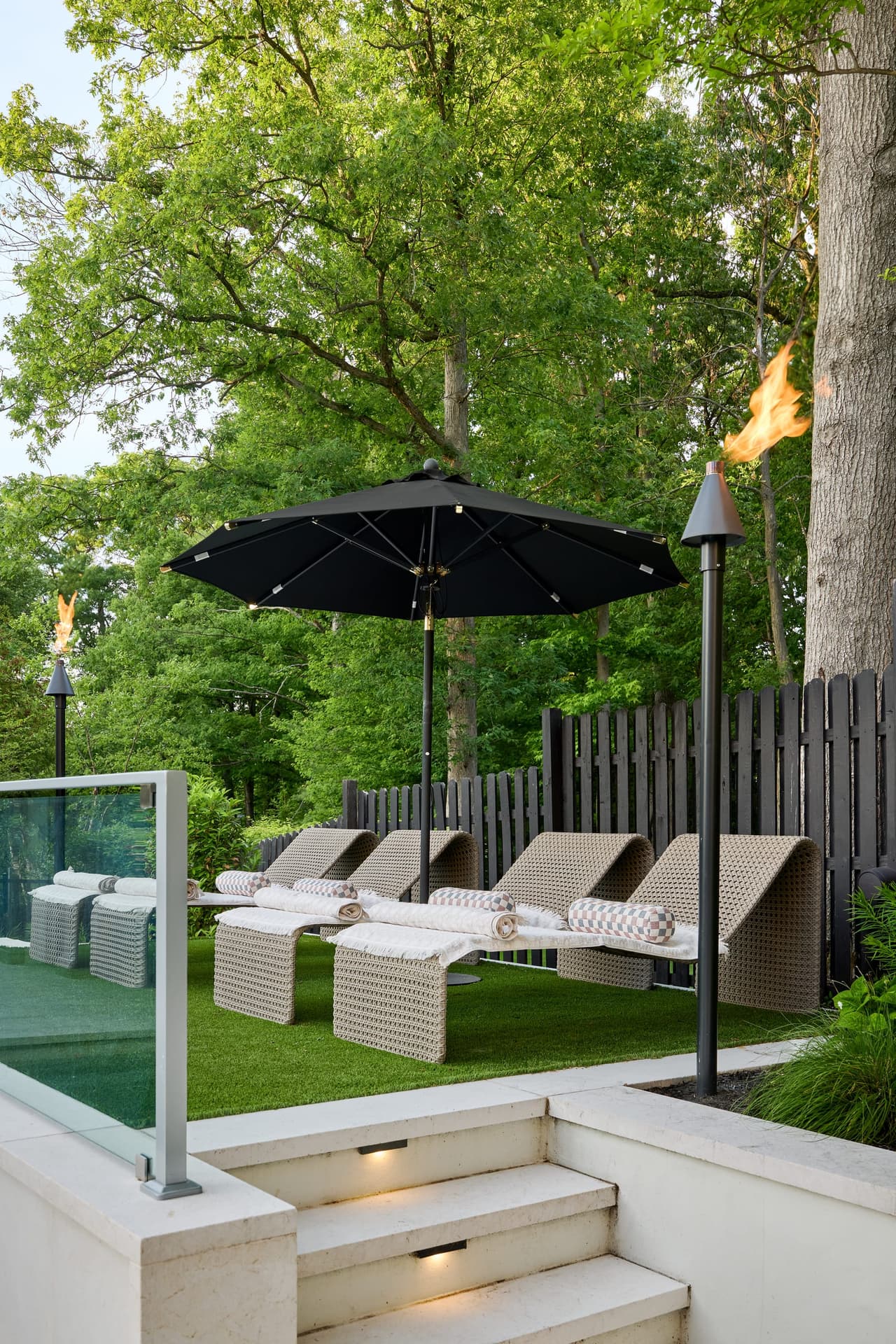 Raised pool terrace at a Westfield, NJ home, with masonry steps built into the wall, a glass railing, woven chaises under an umbrella and a lit fire torch