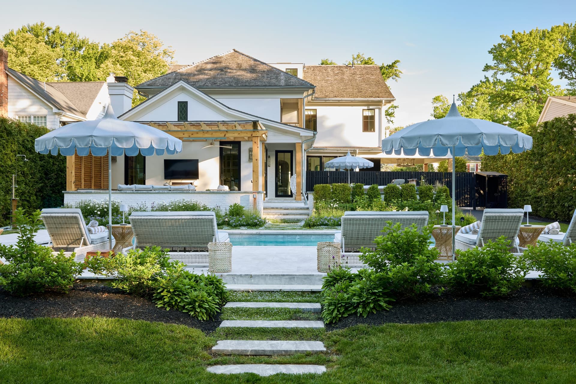 Stone steps rising through the lawn to the pool terrace of a Westfield, NJ new-construction home, with scalloped blue umbrellas, loungers and a covered outdoor room beyond