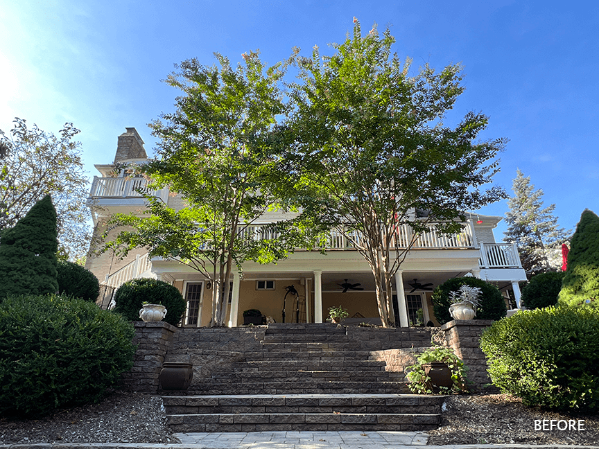 Before photograph of the back of the existing home in Mountainside, NJ, screened by tall trees above a stone terrace, labeled Before in the corner